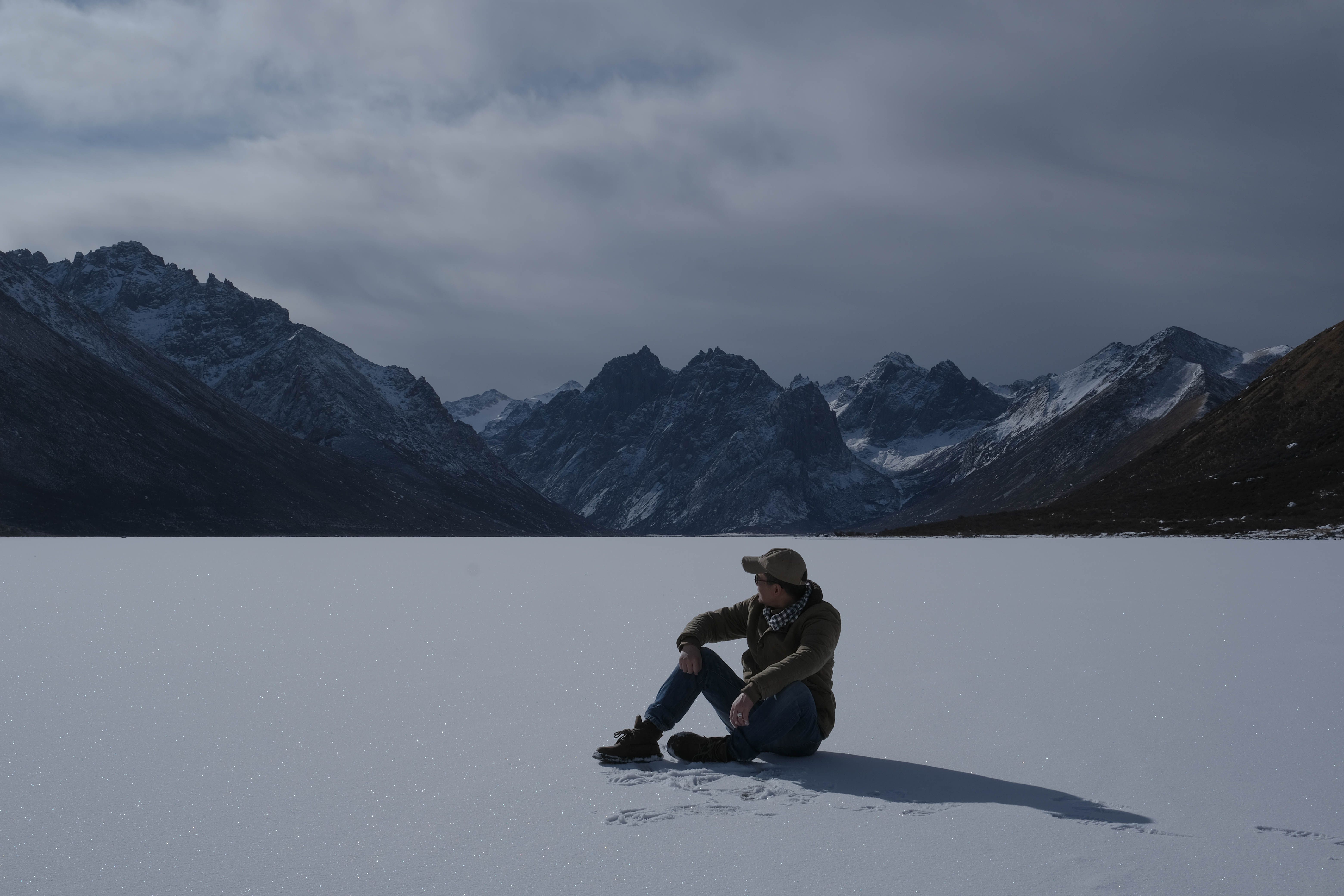 A person sits alone on a frozen lake, facing a vast range of snow-covered mountains.